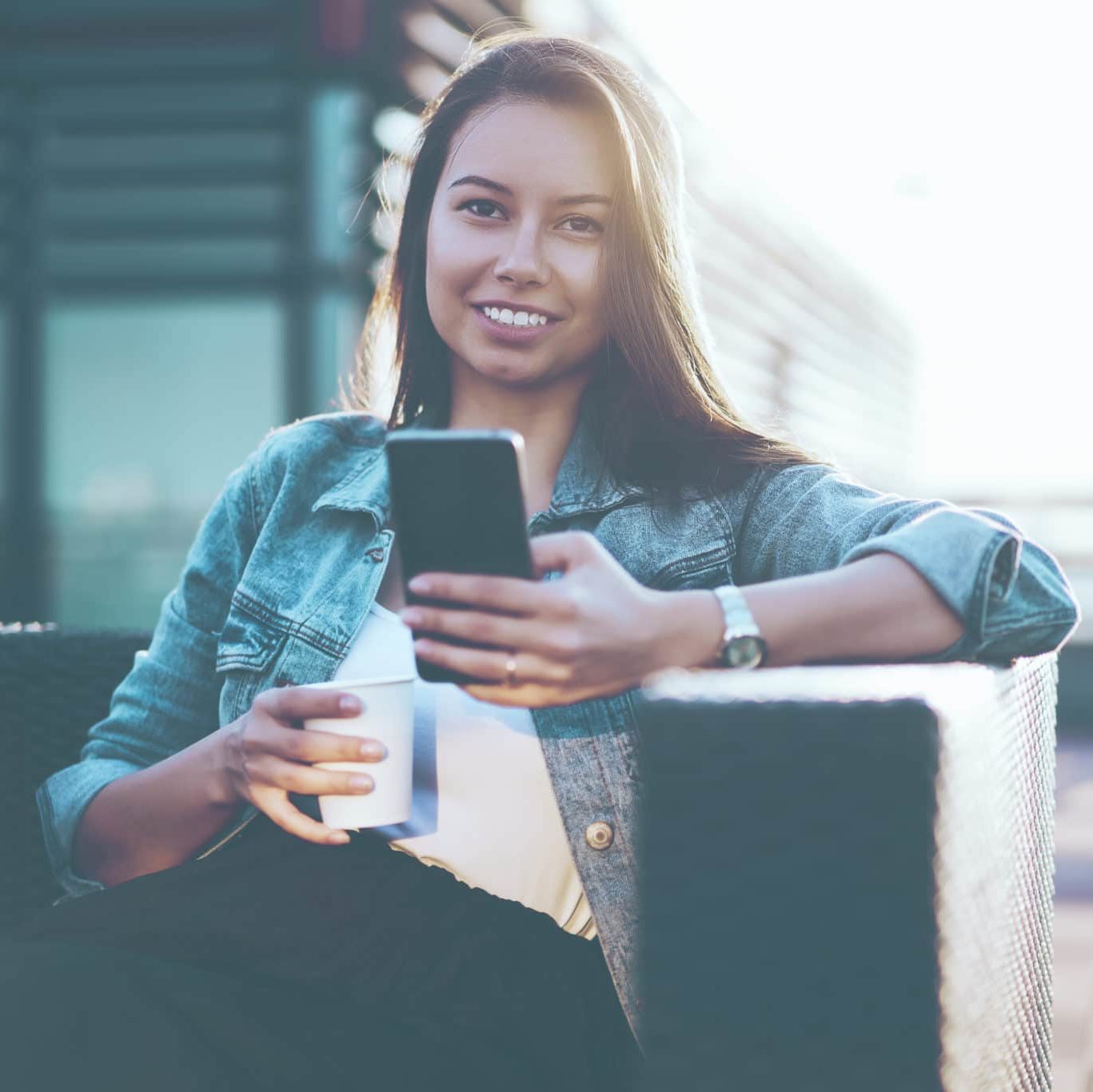 Cheerful hipster girl dressed in denim jacket sending sms message to friend on modern smartphone device.Positive blogger chatting online on mobile phone enjoying tasty coffee sitting outdoors in cafe